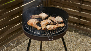 Okrągły grill węglowy na trzech nóżkach z kotletami schabowymi na tle drewnianego ogrodzenia