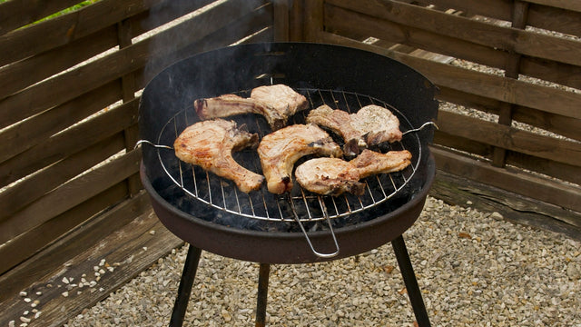 Okrągły grill węglowy na trzech nóżkach z kotletami schabowymi na tle drewnianego ogrodzenia